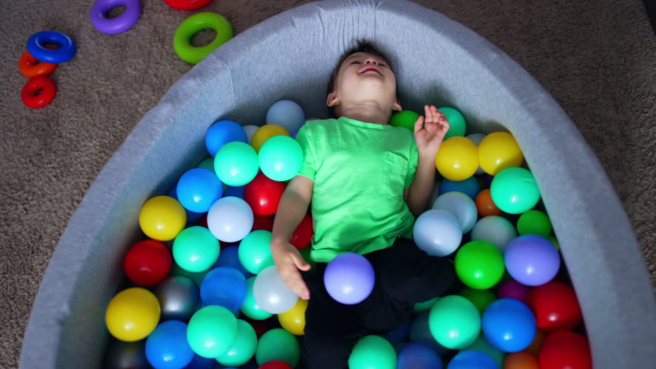 Relaxed Caucasian toddler sitting in the dry pool. Smiling kid lies back on the colorful balls. Top view close up.