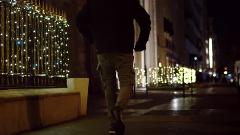 Hombre-Caminando-En-La-Calle-Por-La-Noche-Con-Brillantes-Luces-Navideñas-En-La-Verja-De-Hierro-En-Segundo-Plano