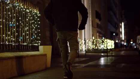 man walking on the street at night with glittering christmas lights on iron fence in background