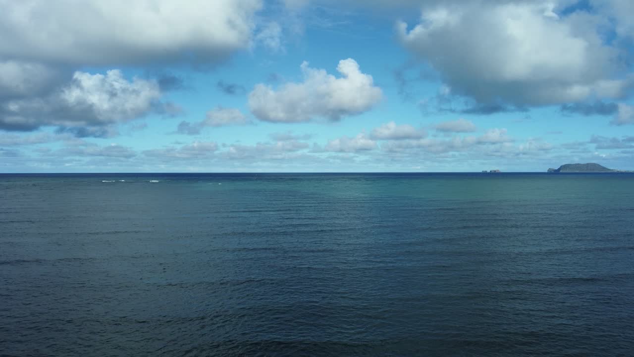 공중 드론 뷰는 하와이 오아후를 둘러싸고 있는 광활하고 푸른 바다의 물을 포착하며, 은 하늘 아래 황금색 해안선과 멀리 떨어진 섬들을 향해 부드러운 파도를 드러니다.