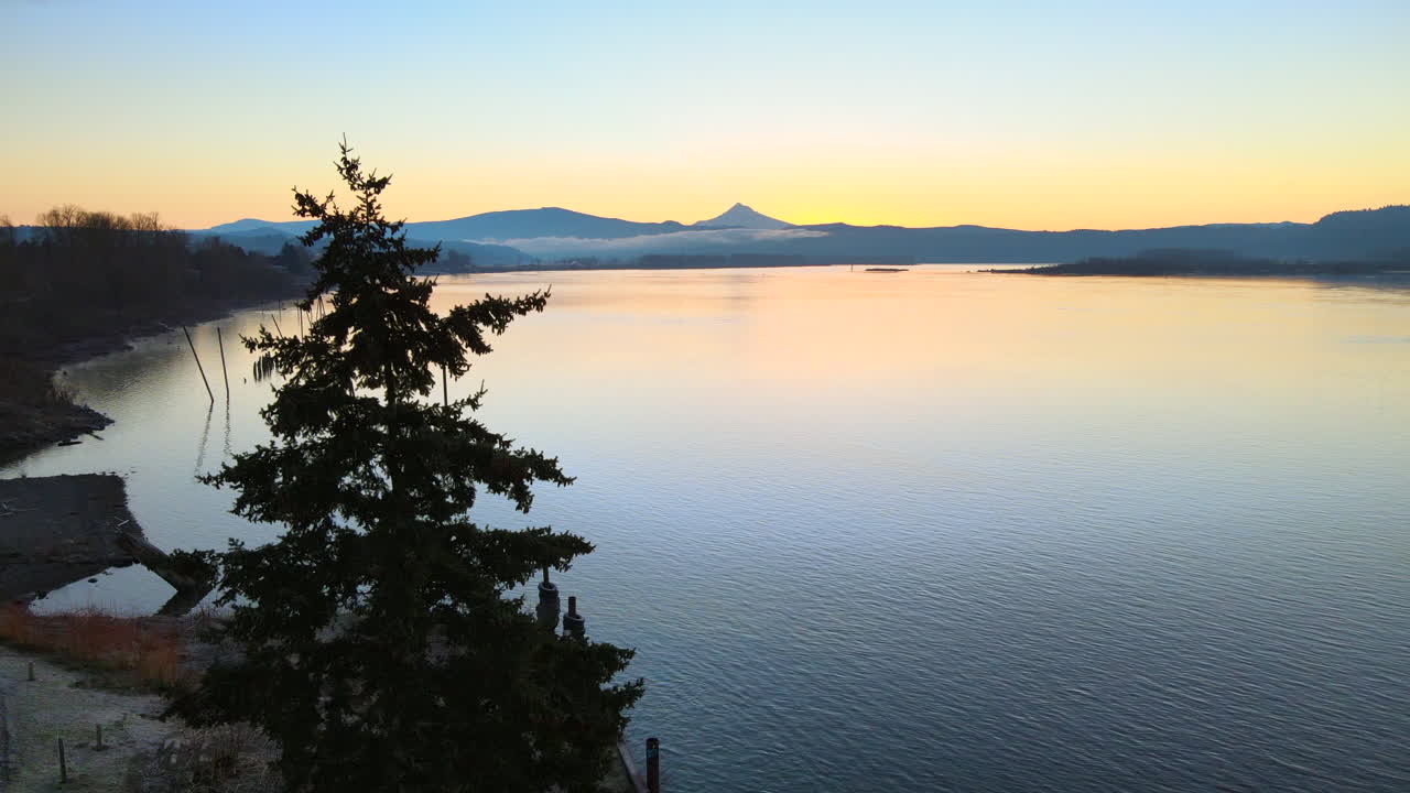 ドローンが飛行し、木々の上に上昇し、美しい日の出の間に大きな湖の素晴らしい景色を明らかにし、山を背景にしています