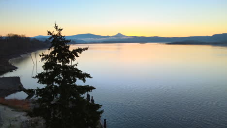 drone taking flight, rising above the trees and revealing a stunning scenery of a large lake during a beautiful sunrise, mountains in background