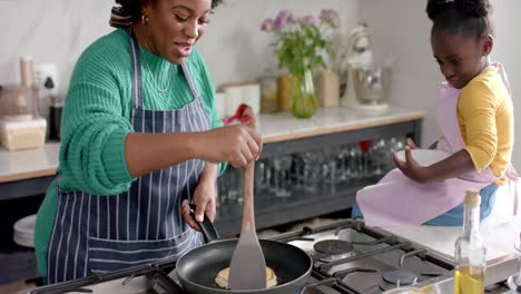 happy african american mother and daughter frying pancakes in kitchen, slow motion