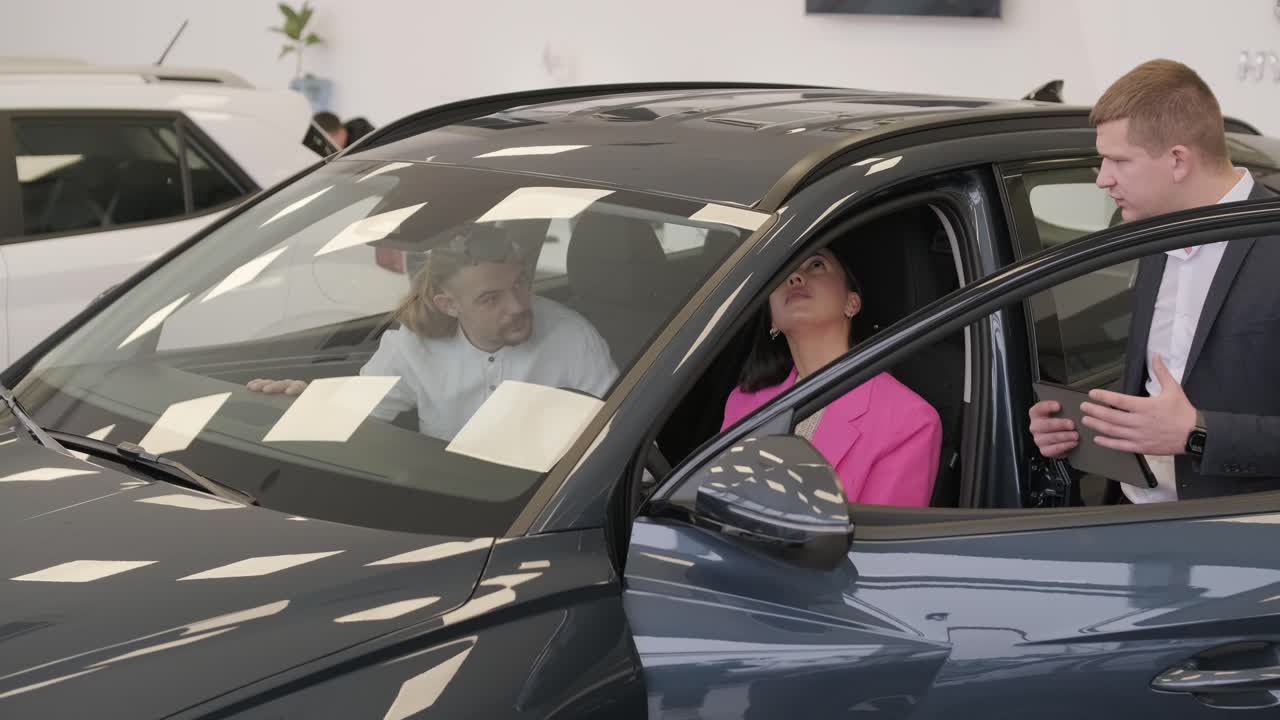 un joven vendedor de coches mostrando a una pareja de jóvenes un nuevo automóvil en el salón de concesionarios.