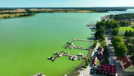 Aerial-view-of-a-vibrant-lakeside-town-with-boats-docked-at-the-marina,-lush-greenery,-and-fields-in-the-distance,-under-a-clear-blue-sky