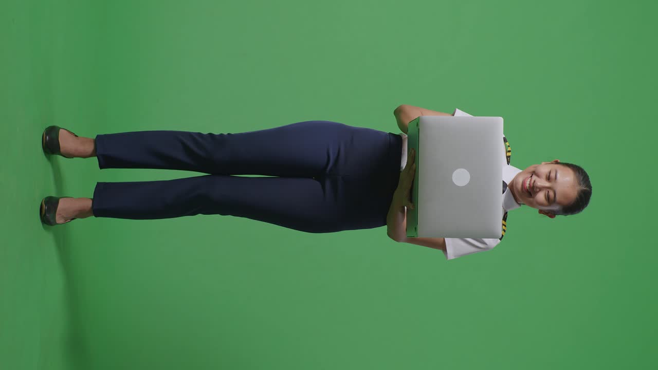 cuerpo lleno de piloto mujer asiática sonriendo y usando una computadora portátil mientras está de pie en el estudio de fondo de la pantalla verde