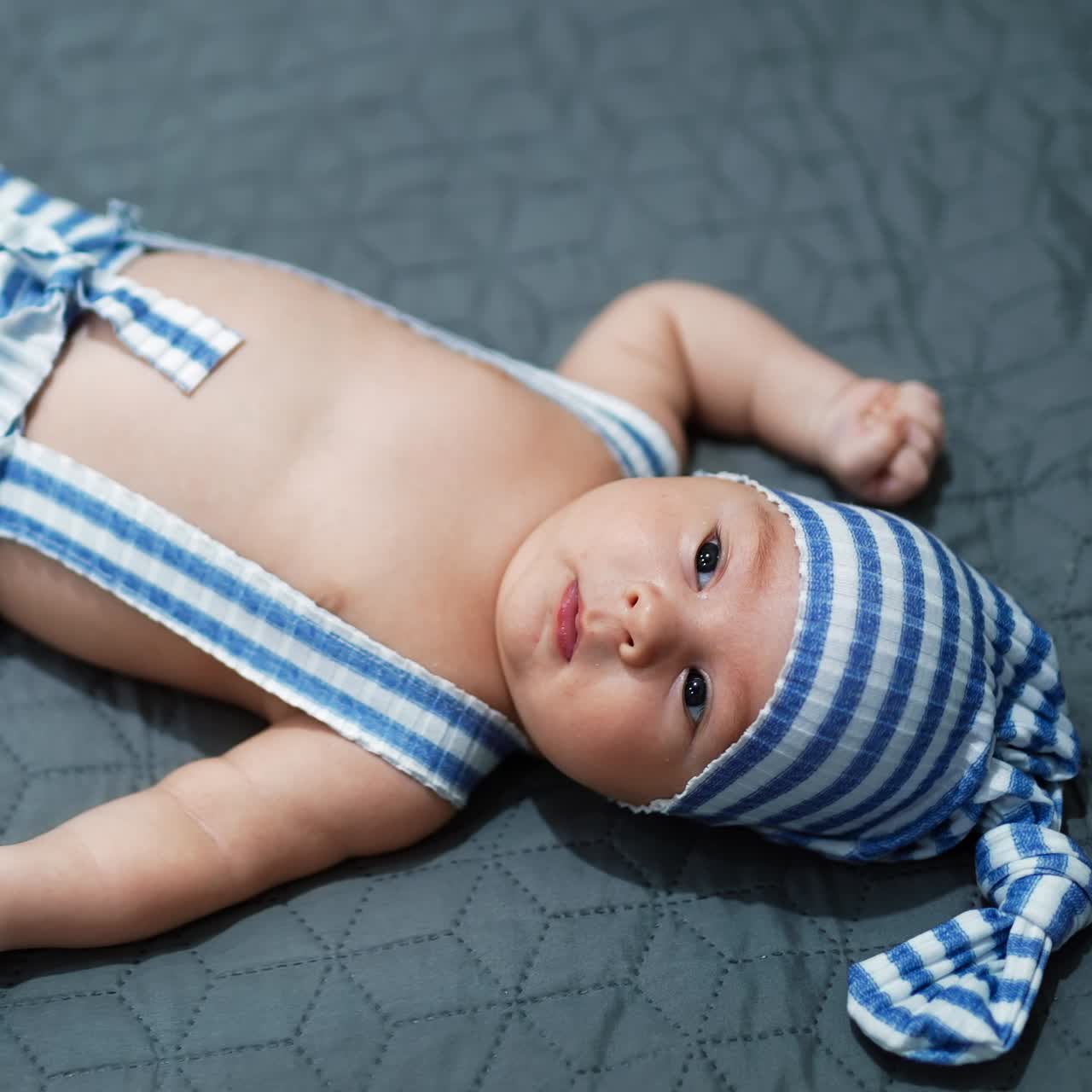 Little boy in a funny striped costume lying calmly on the bed. Baby with naked belly resting on grey background. Close up