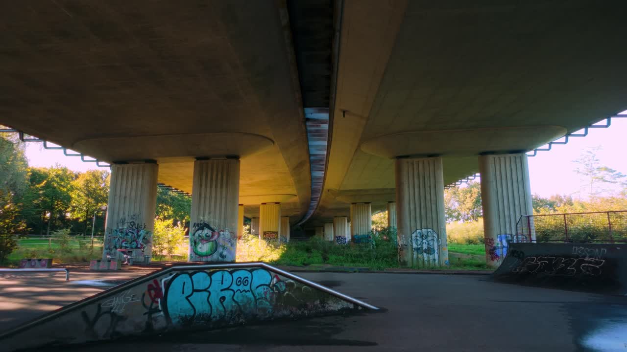 Wide-angle shot of urban skate park beneath highway overpass. Concrete structure is adorned with graffiti, and small ramps are visible. Sun shines through surrounding trees, creating atmospheric scene