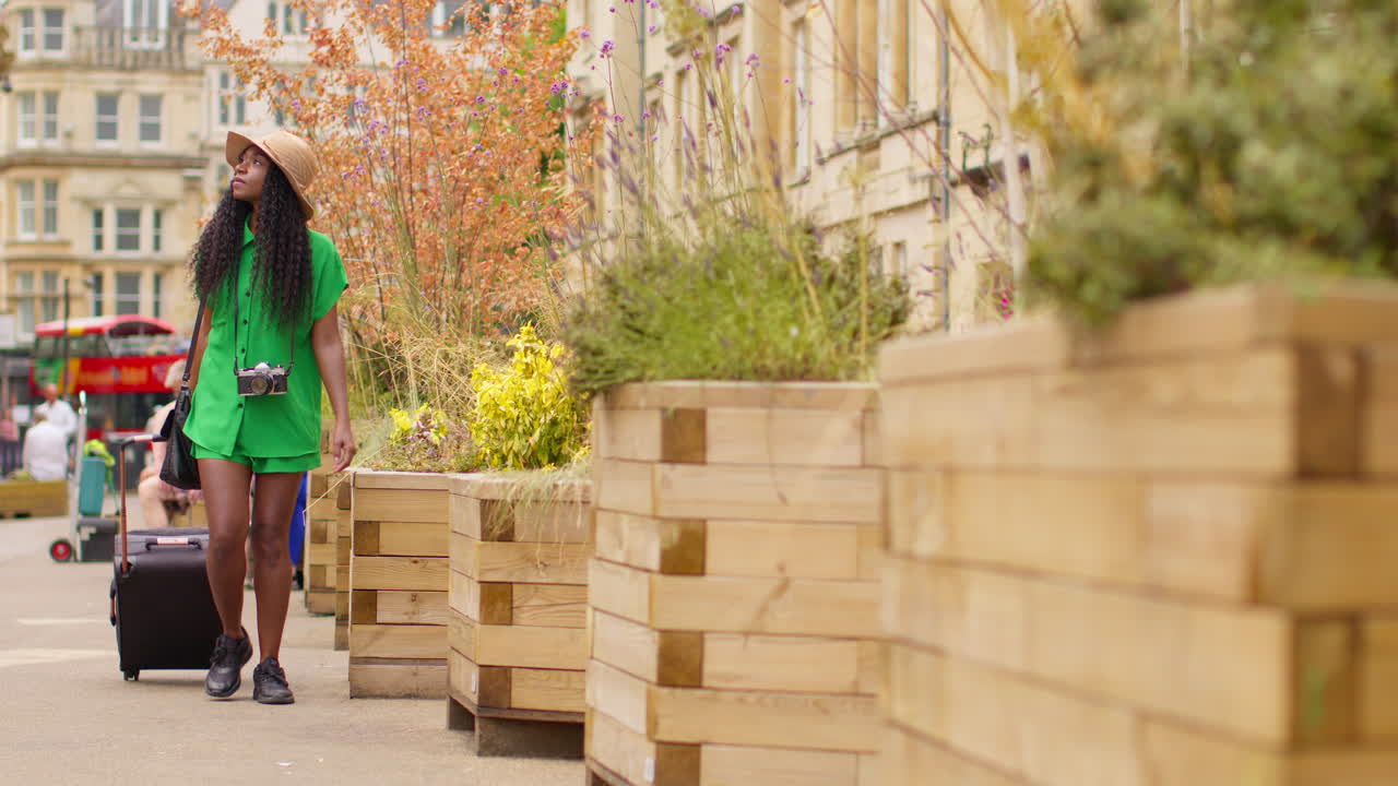 영국 옥스퍼드에서 휴가를 보내고 있는 카메라를 든 여성 관광객이 바가 있는 가방을 끌고 브로드 스트리트를 따라 고 있습니다.