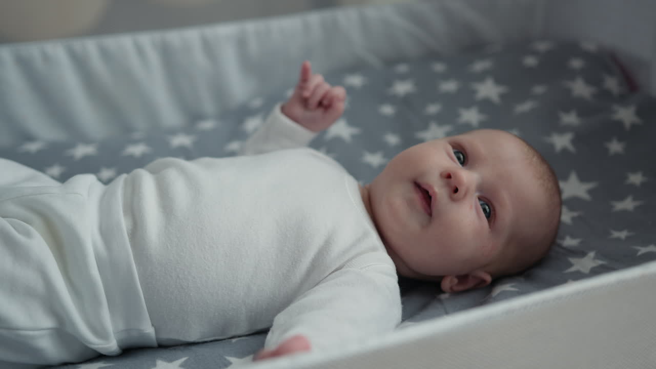 Neonato in tutina bianca sdraiato sulla schiena nella culla con lenzuola a stelle, arredamento per la cameretta del bambino.