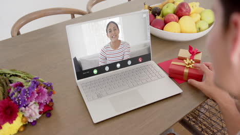mujer caucásica sosteniendo un regalo rojo usando una computadora portátil con una mujer biracial en la pantalla