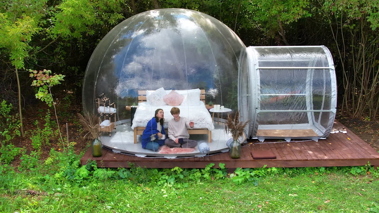 Et par sidder i et gennemsigtigt boble-telt ved glamping. De sidder under et tæppe, holder kopper og snakker sammen. Set fra en drone med en frodig skov i baggrunden.
