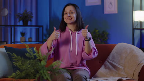 Portrait-of-happy-woman-sitting-on-sofa-at-home-raises-thumbs-up,-likes-good,-agrees-with-positive