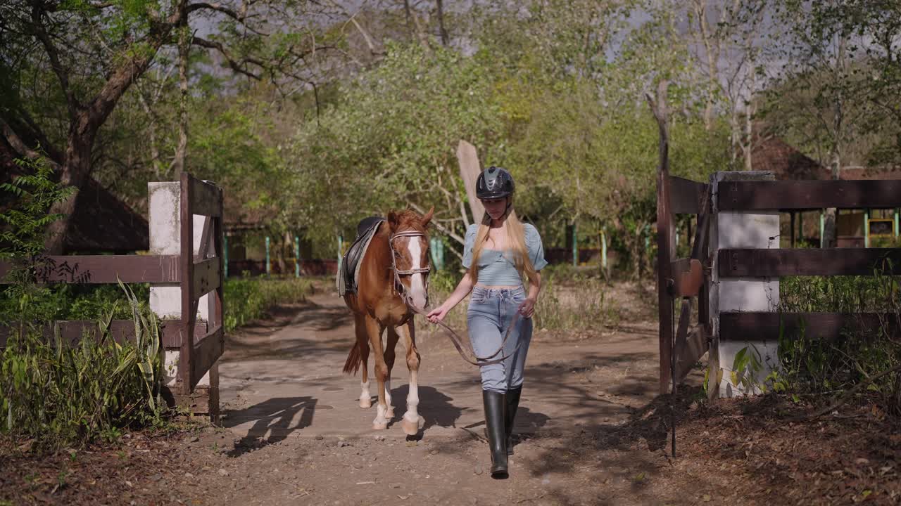 Una donna cammina con calma conducendo un cavallo marrone, indossando un casco e dei jeans. La scena cattura fiducia, controllo e connessione tra uomo e animale in un ambiente naturale all'aperto