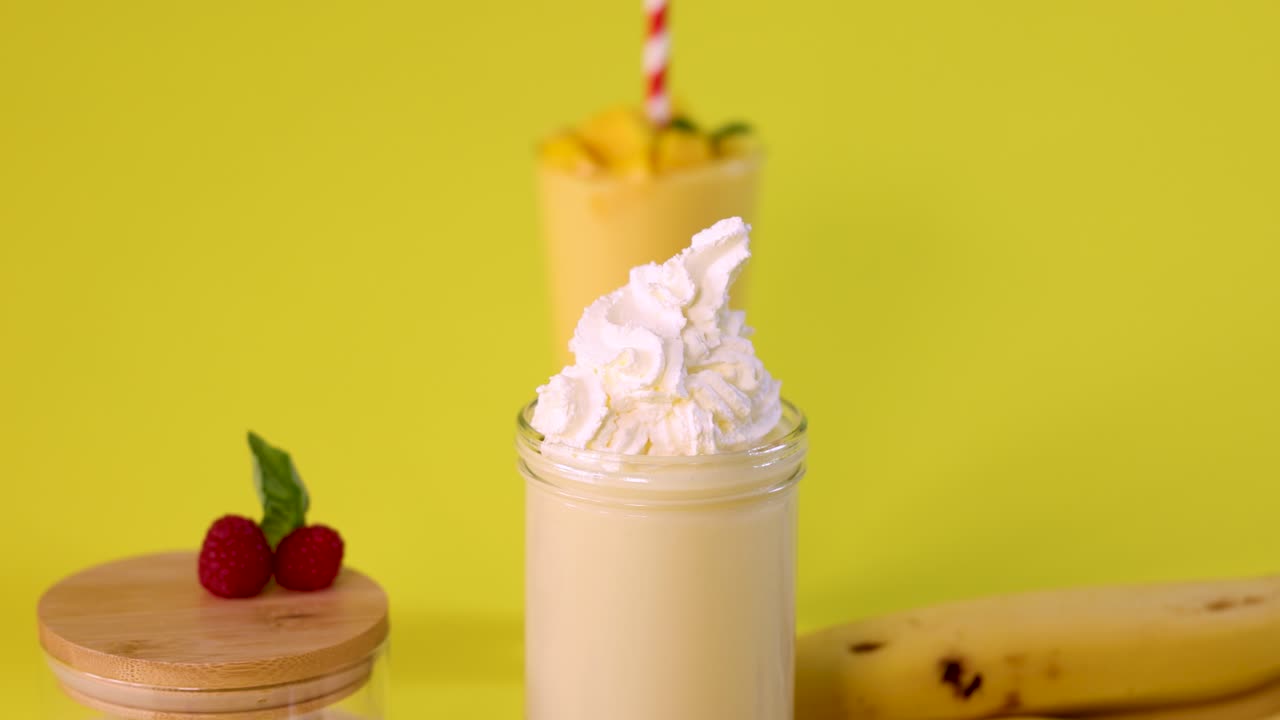 Hand tops banana milkshake with whipped cream, bright yellow background, vibrant lighting, static camera