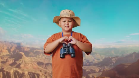 asian boy with a hat and binoculars making heart shape gesture while traveling at the top of mountain. boy researcher examines something, travel tourism adventure concept