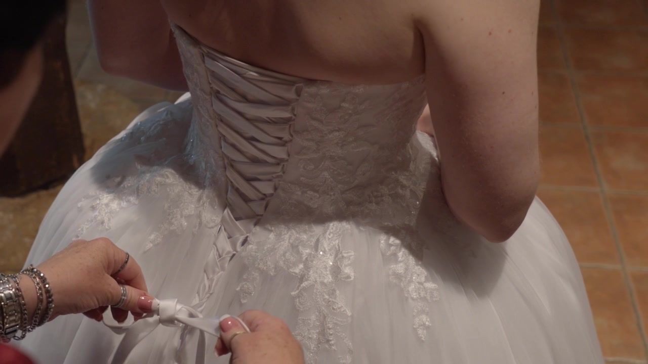 Hands tying the lace back of a white wedding dress on tiled indoor floor