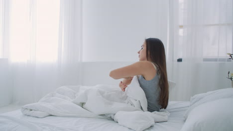 beautiful brunette slowly waking up in the morning stretches and gets up from the bed sun shines on her from the big window. happy young girl greets new day