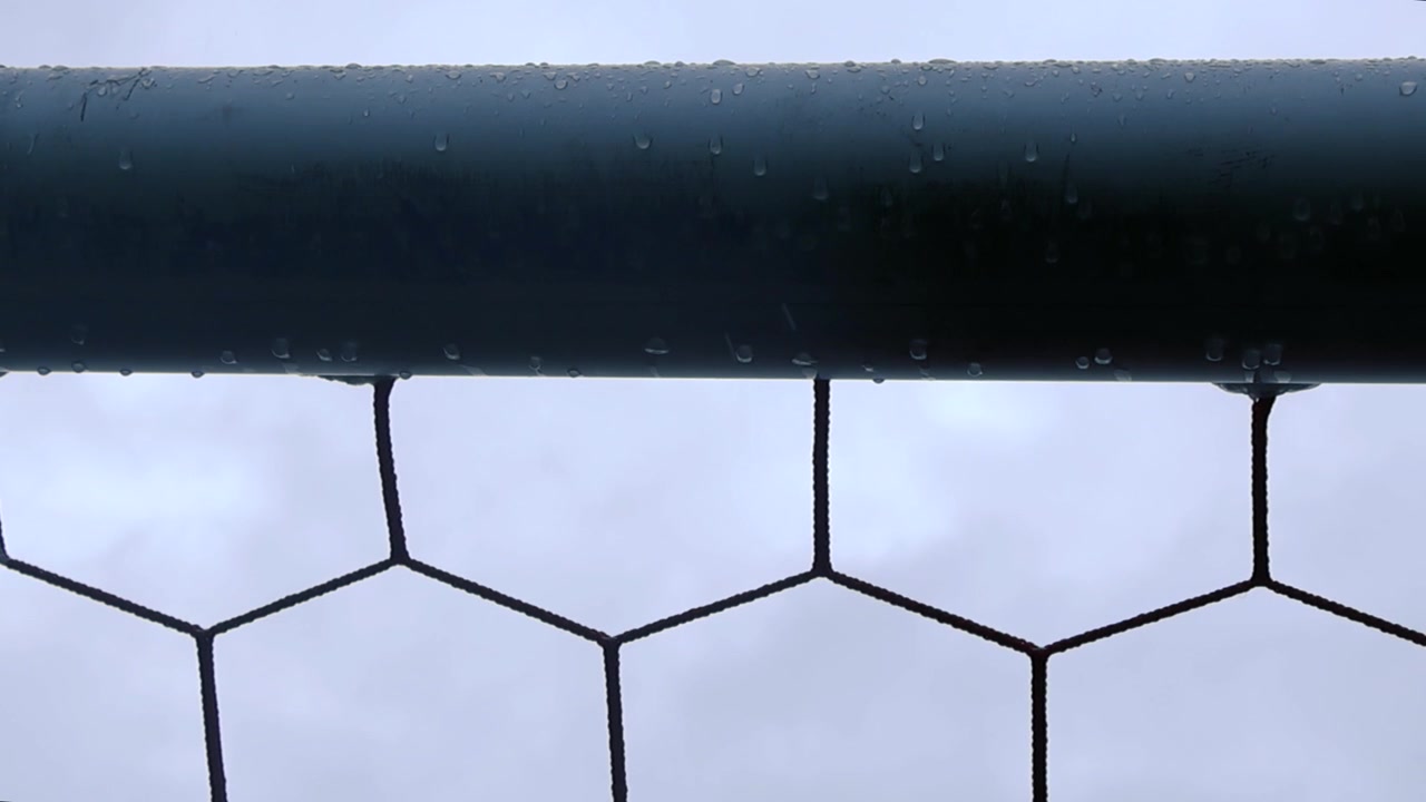 Nærbilde av en våt fotballmålramme og sekskantet nett på en regnværsdag, med regndråper som faller mot en grå og skyet himmel, som symboliserer en utsatt eller avlyst kamp.