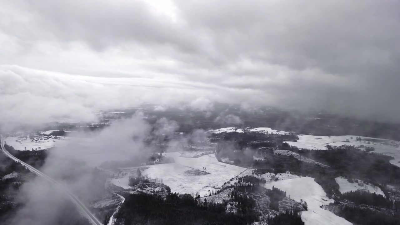 Tjocka grå moln driver över ett lapptäcke av snötäckta fält och skog medan en drönare glider över ett lugnt lantligt landskap
