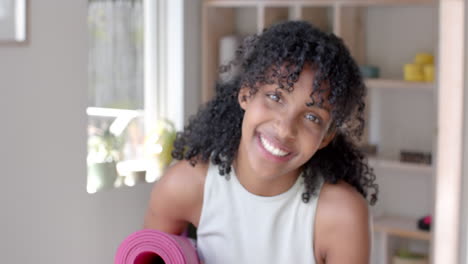 Portrait-of-happy-biracial-fitness-teenage-girl-holding-yoga-mat-in-white-room,-slow-motion