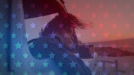 Animation-of-American-flag-waving-over-mixed-race-woman-in-car-looking-out-of-window-by-seaside