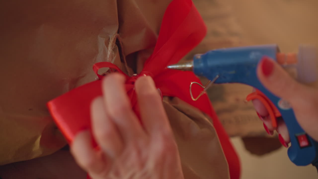 Primo piano di mani che incartano un regalo di Natale con un fiocco di raso rosso, applicando colla a caldo per fissare il nodo su carta kraft marrone, manicure accurata visibile, illuminazione ambientale calda, atmosfera accogliente di artigianato natalizio.