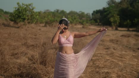 mujer en sari rosa posando con gracia en un campo seco, luz natural del día