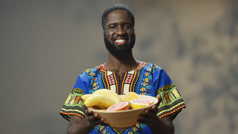 portrait of young cheerful african american man in traditional clothes holding and showing a plate with tropical fruit while looking at camera