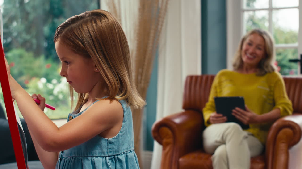 딸과 함께 가족은 소파에서 디지털 태블릿 시계를 가진 어머니로 easel에 그림을 그리고 있습니다.