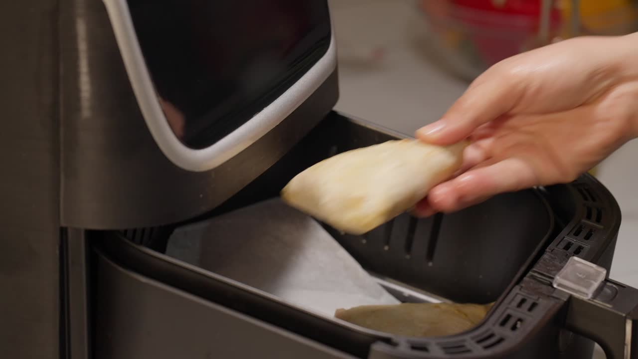 Un primo piano di una mano che mette un alimento congelato o preparato (probabilmente un involtino primavera, un bastoncino di mozzarella o qualcosa di simile) nel cestello di una friggitrice ad aria nera