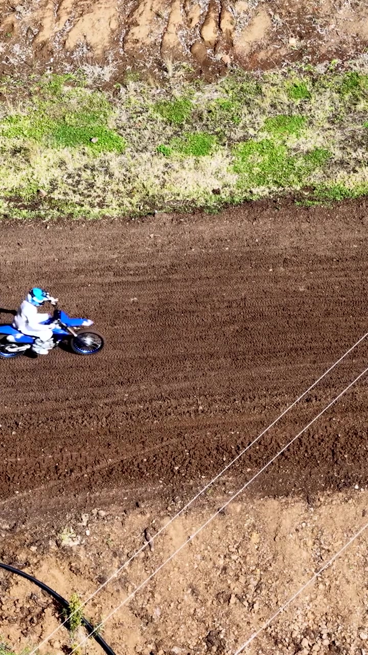 Uma tomada de drone em ângulo elevado mostra um piloto de motocross acelerando por uma pista de terra marrom.