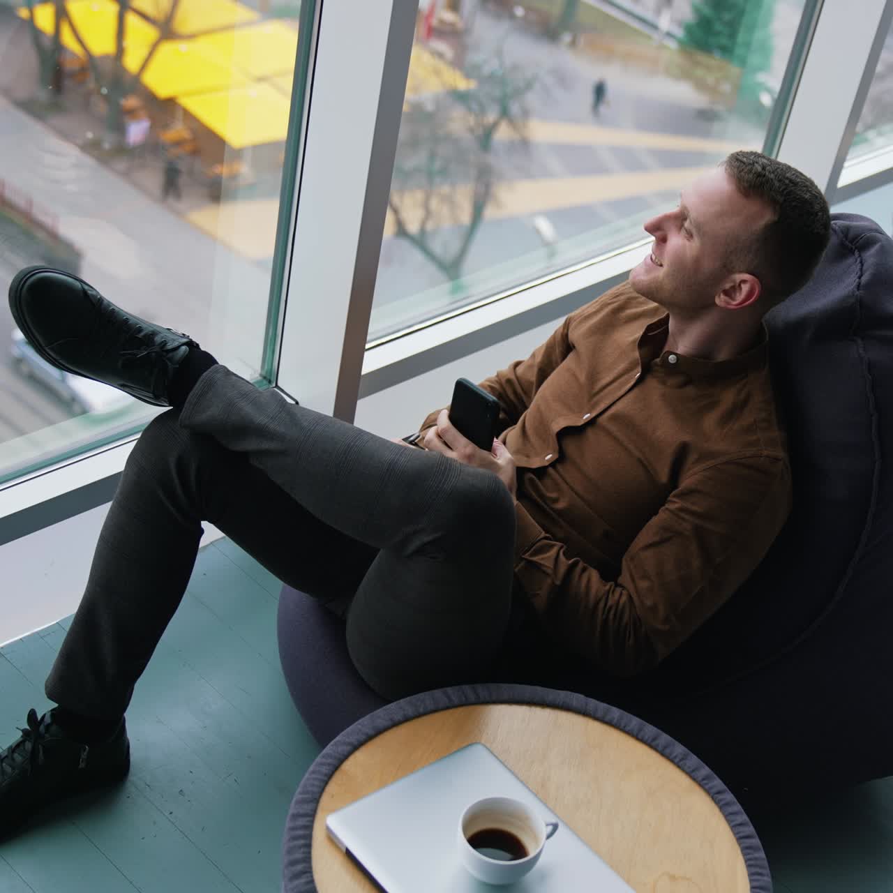 Businessman relaxing on view from office window. Guy with a phone looking at city through window on comfortable ottoman. Work life balance concept. Top view