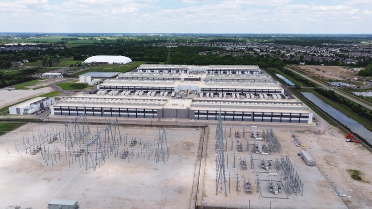 Fremadrettet dronebillede over et hyperscale-datacenter med store batterier og køleenheder nær et beboelseskvarter