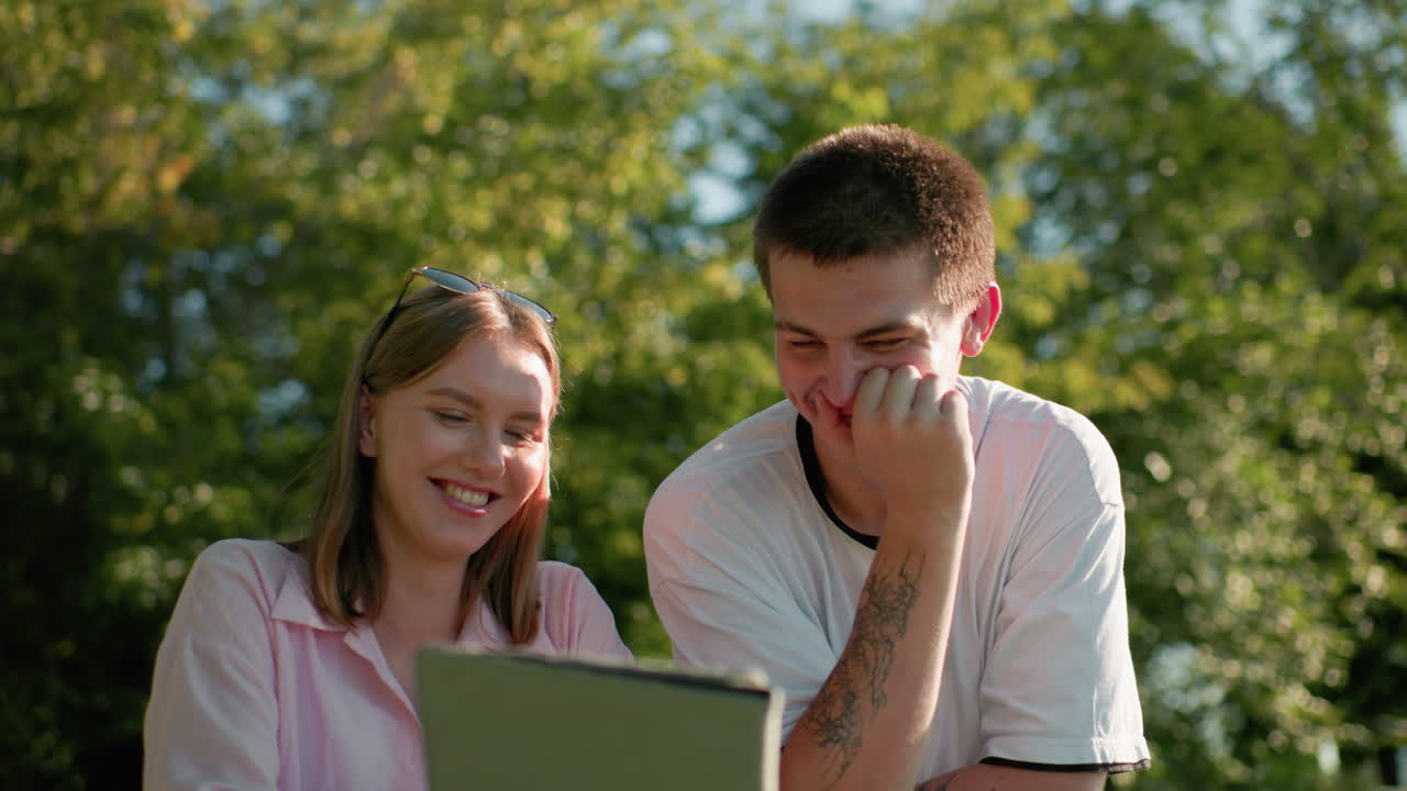mujer de top rosa sonriendo mientras le muestra a su hermano algo en su tableta, asintiendo con la cabeza y riendo, mientras él señala la tableta con una mano y se cubre la boca con la otra