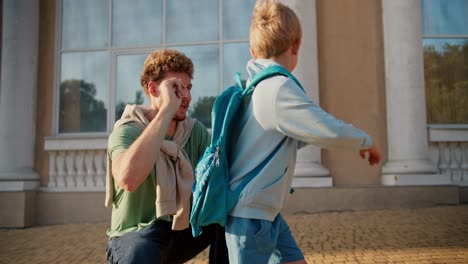 a father with curly hair in a green t-shirt sends his little blond son in a blue jacket and with a turquoise backpack to school. little elementary school student runs to school