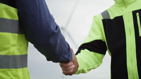 part of two caucasian and latin engineers standing on wind turbine field and shaking hands.