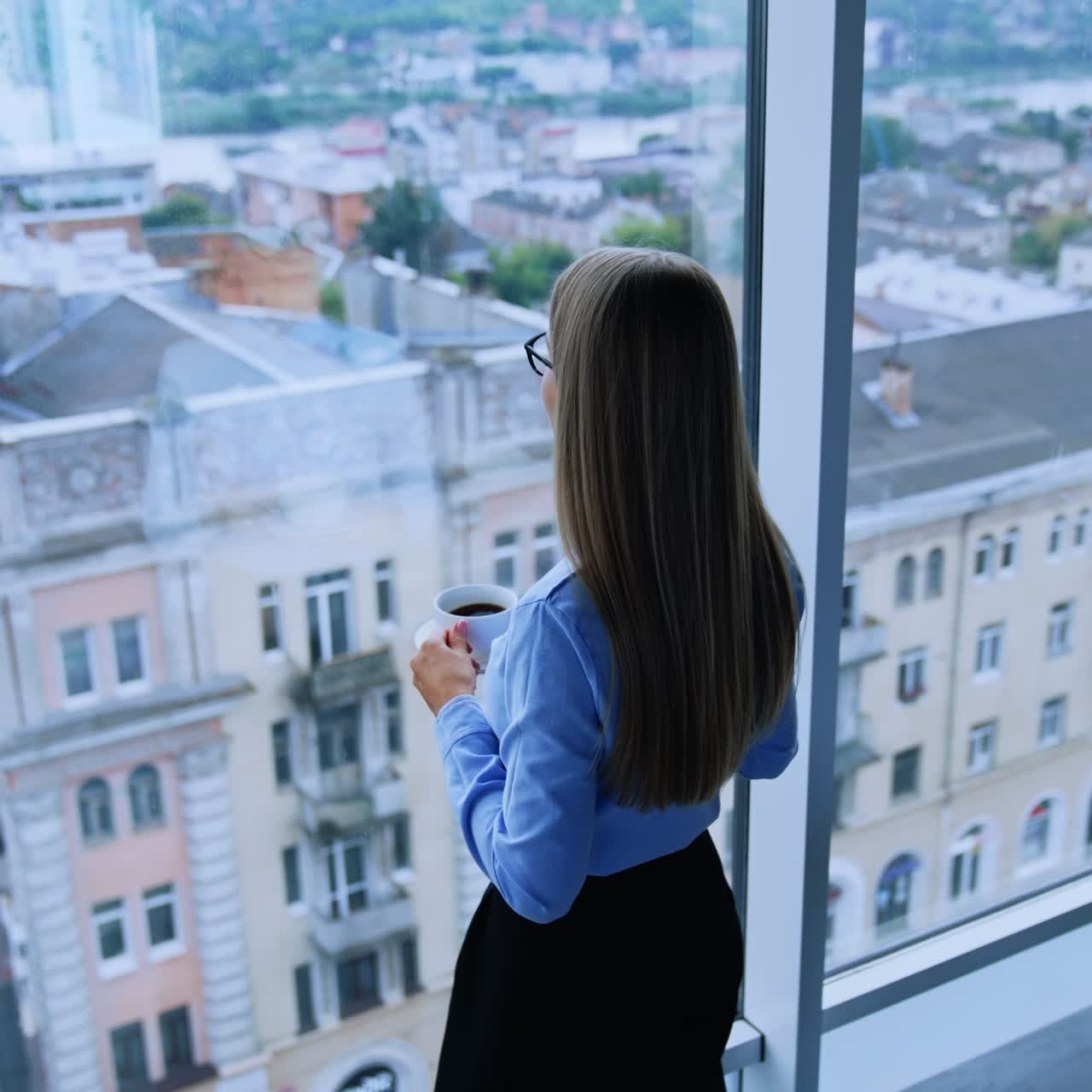 Lunch break in the modern office with beautiful view from window. Blonde female employee walking up to window with a cup of coffee. High angle view