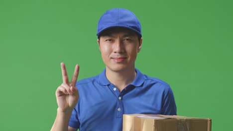 푸른 유니폼을 입은 아시아 남성 택배사의 클로즈업은 스튜디오에서 녹색 스크린 배경에 상자를 배달하는 동안 몸짓 평화와 미소를 짓고 있습니다.