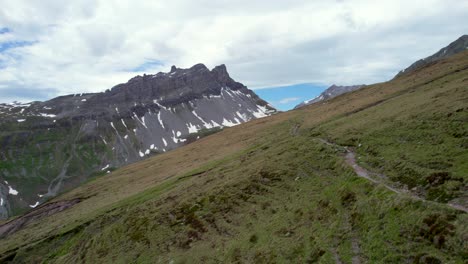 Aerial-drone-footage-slowly-reversing-and-rising-to-reveal-a-hiking-trail-leading-along-grassy-slopes-towards-a-dramatic,-jagged-mountain-ridge-with-residual-patches-of-snow