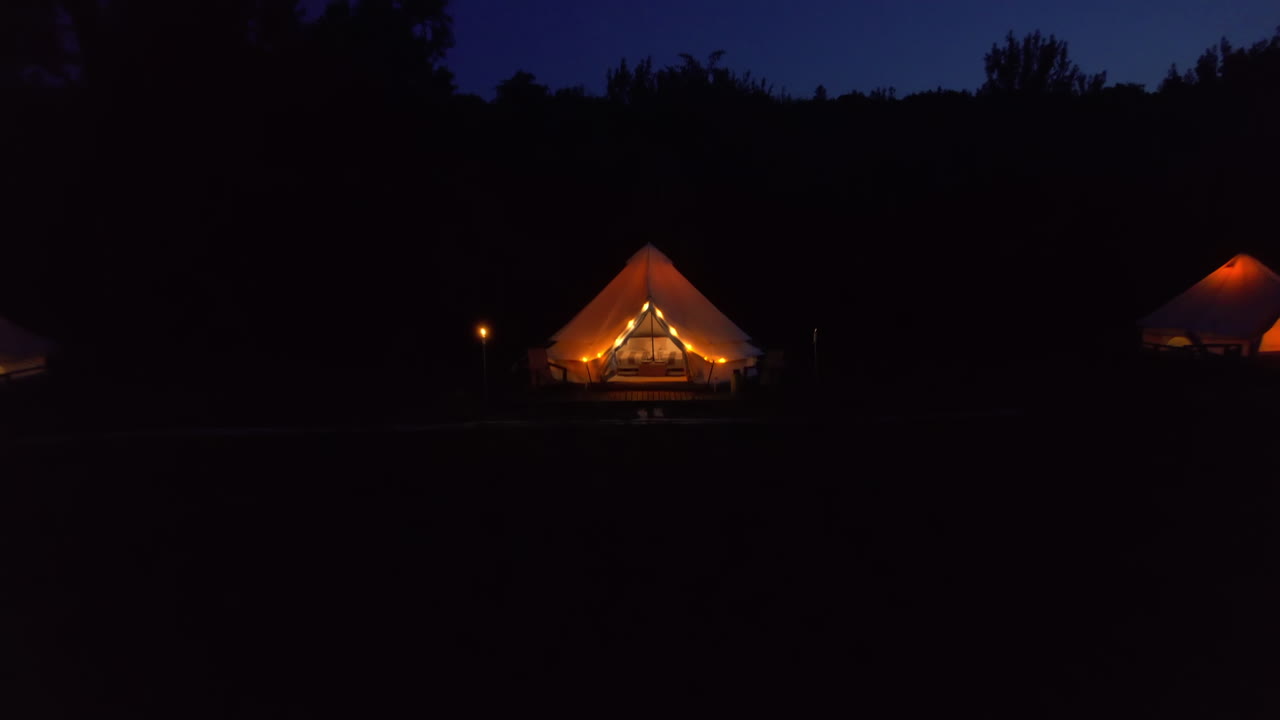 Vista aerea dall’alto di interni ed esterni di una tenda con letto e illuminazione in un glamping, circondata da una foresta di notte