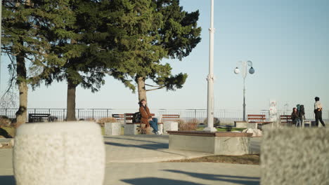 a woman wearing a brown coat, blue jeans, and white shoes sits alone on a bench in an urban park, her black backpack beside her. she appears to be deep in thought, with a depressed demeanor