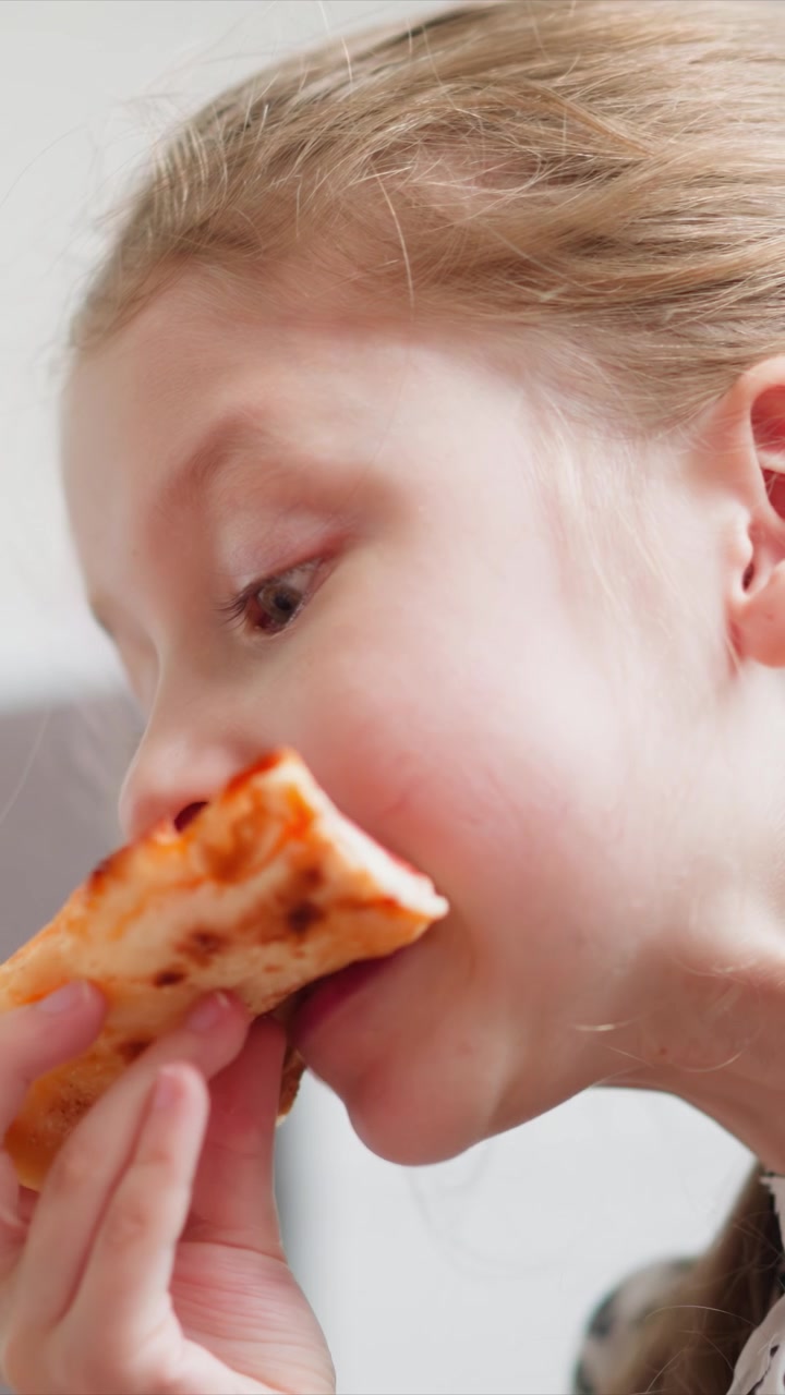 Close up of young girl eating pizza. child holds and takes bites from food with soft lighting highlighting her profile against indoor background. Details include visible texture on both