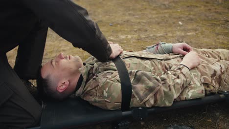 Close-up-a-confident-man-military-man-in-dark-green-army-clothes-fastens-a-wounded-soldier-in-camouflage-clothing-on-a-stretcher-with-a-belt-in-combat-conditions-in-the-steppe