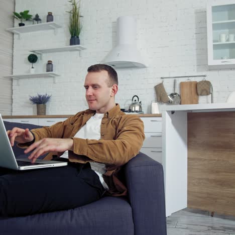 young man working at home. relaxed man laying on a sofa and typing on a laptop. student studying online at home during quarantine. remote job