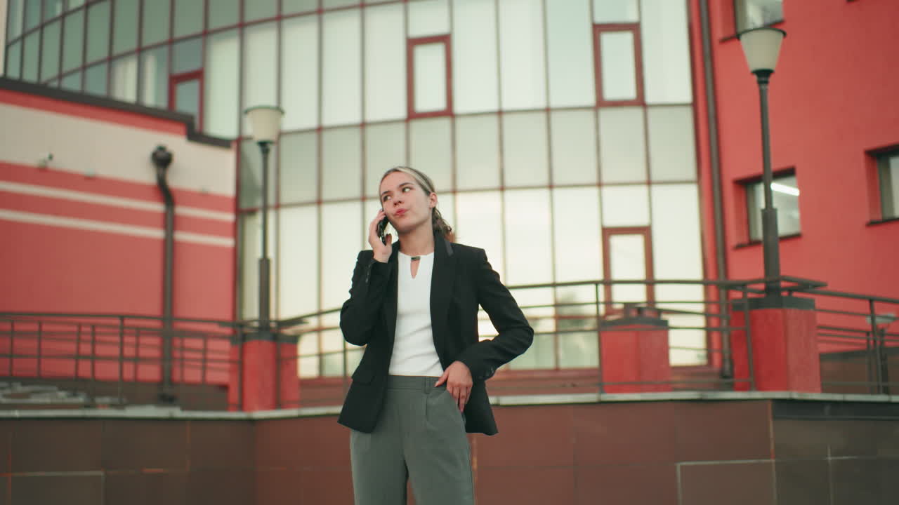 Lady in professional outfit standing with tired expression while on phone call in urban environment, hand in pocket, modern building with large windows in background