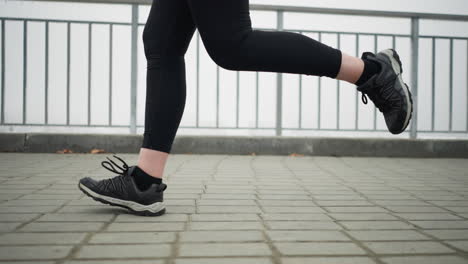 leg view of lady in black sneakers and leggings jogging near iron railing of bridge with partial view of foggy cityscape during winter