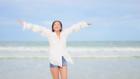 woman with white shirt and shorts standing on seashore turns on herself raises arms up to blue sky