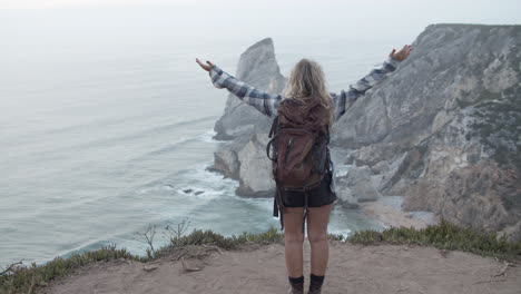 Woman-is-spreading-her-arms-on-top-of-a-cliff-looking-over-the-sea,-majestic-landscape-view-over-the-beach-and-water