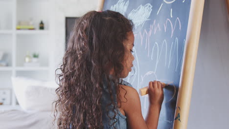 young hispanic girl drawing with chalks at a blackboard at home, close up, back view